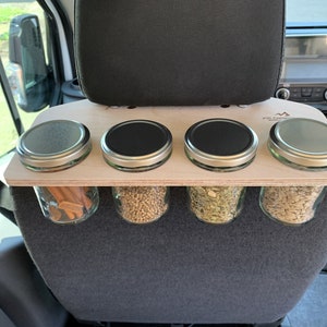 May include: A wooden shelf with four glass jars with lids, mounted on the back of a car seat. The jars contain various spices and ingredients. The shelf is labeled "Fit Campers".