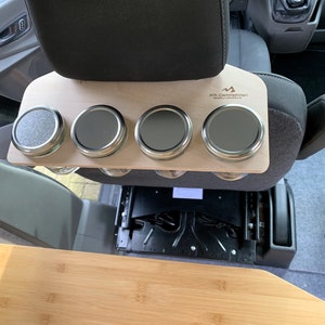 May include: A wooden shelf with four metal jars attached to it. The shelf is mounted on the back of a car seat. The shelf is labeled "FB Campervan".