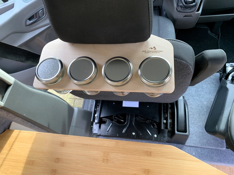 May include: A wooden shelf with four metal jars attached to it. The shelf is mounted on the back of a car seat. The shelf is labeled "FB Campervan".