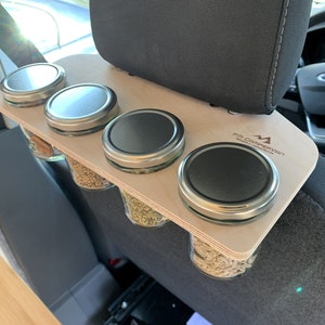 May include: A wooden organizer with four glass jars with lids attached to the headrest of a car seat. The organizer has the text "FM Campervan" printed on it.