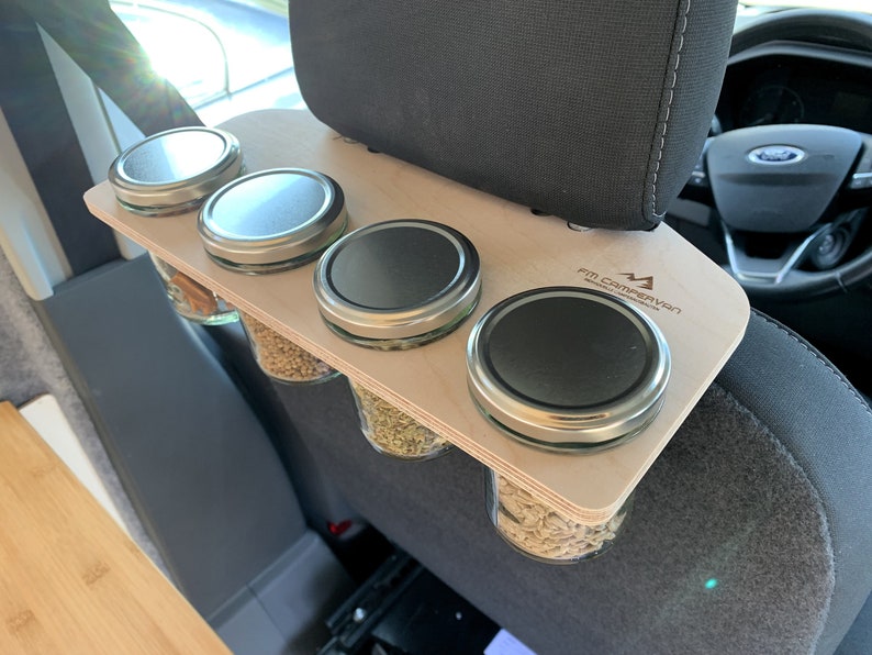 May include: A wooden organizer with four glass jars with lids attached to the headrest of a car seat. The organizer has the text "FM Campervan" printed on it.