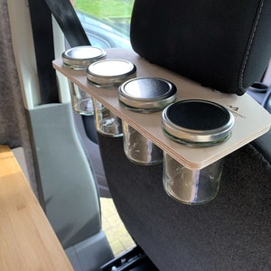 May include: A wooden spice rack with four glass jars with metal lids attached to the headrest of a car seat. The rack is made of light wood and has the text "Adventure Awaits" printed on it.