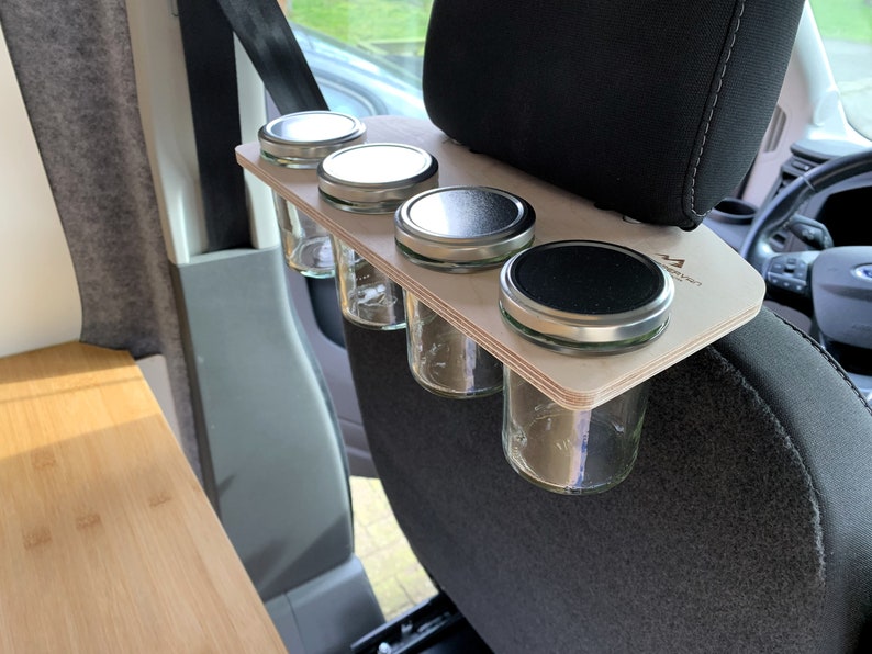 May include: A wooden spice rack with four glass jars with metal lids attached to the headrest of a car seat. The rack is made of light wood and has the text "Adventure Awaits" printed on it.