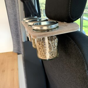 May include: A wooden shelf with four glass jars filled with sunflower seeds, attached to the headrest of a car seat.