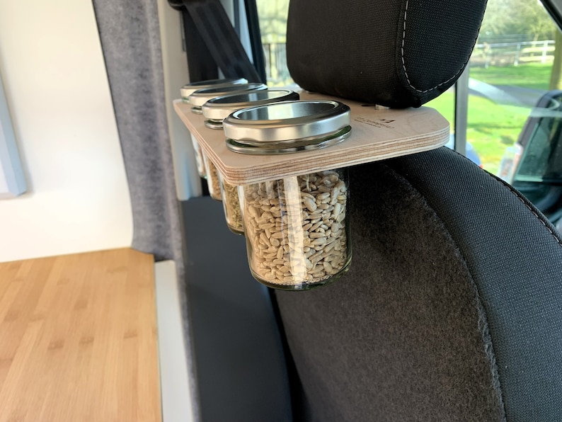 May include: A wooden shelf with four glass jars filled with sunflower seeds, attached to the headrest of a car seat.