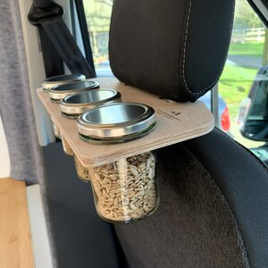 May include: A wooden shelf with four glass jars with metal lids, filled with sunflower seeds, attached to the headrest of a car seat. The shelf is labeled "The Camper Co."