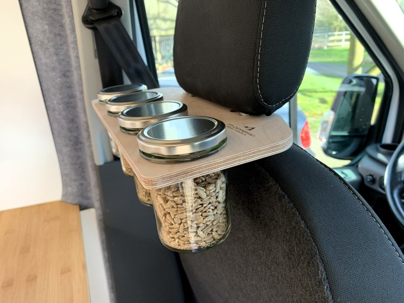 May include: A wooden shelf with four glass jars with metal lids, filled with sunflower seeds, attached to the headrest of a car seat. The shelf is labeled "The Camper Co."