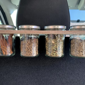 May include: Four glass jars with silver lids are mounted on a wooden shelf. The jars contain various spices and ingredients, including cinnamon sticks, coriander seeds, dried herbs, and sunflower seeds.