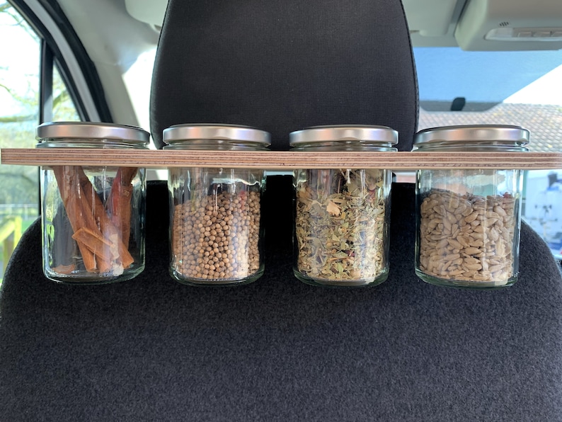 May include: Four glass jars with silver lids are mounted on a wooden shelf. The jars contain various spices and ingredients, including cinnamon sticks, coriander seeds, dried herbs, and sunflower seeds.