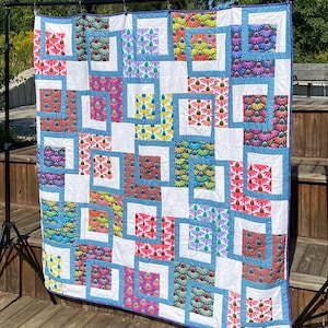 May include: A colorful handmade quilt with a patchwork design. The quilt features squares with floral patterns in various colors, including red, pink, yellow, and blue, set against a white background. The quilt is displayed outdoors, hanging on a black stand.