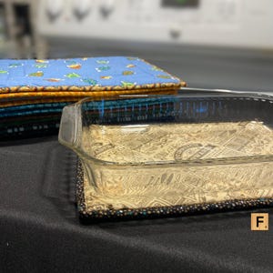 May include: A glass baking dish sits on a brown and black patterned fabric hot pad. The hot pad has a black border. A stack of blue, orange, and green fabric squares is in the background.