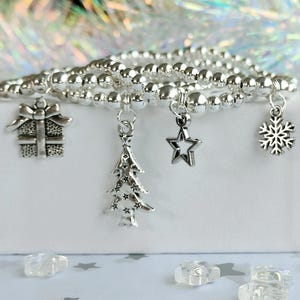 May include: Silver beaded bracelets with festive charms: a gift, Christmas tree, star, and snowflake. The bracelets are presented on a white surface with a shimmering background, ideal for the holiday season.