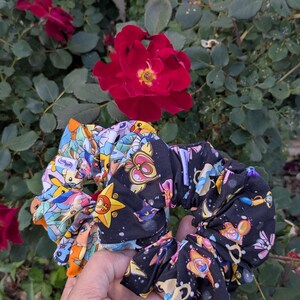 May include: Two colorful scrunchies with cartoon character prints. One features a black background with a celestial design, and the other has a light blue background with various characters. Red roses and green foliage are in the background.