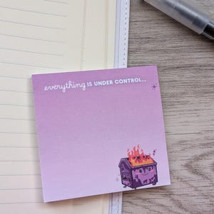 May include: A square purple sticky note featuring a gradient design with the text "everything IS UNDER CONTROL..." and a graphic of a smiling dumpster on fire. The note is placed on a lined notebook page next to a clear plastic pen.