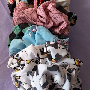 May include: A collection of colorful scrunchies with various patterns. The scrunchies feature designs such as geometric shapes, anime characters, and floral prints. The colors include pink, blue, black, white, and cream. These hair accessories are made of fabric.