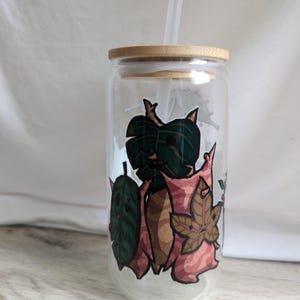 May include: A clear glass reusable drinking cup with a bamboo lid and a clear plastic straw. The cup has a colorful design of leaves and vines on the side.