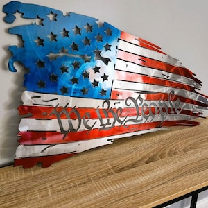 May include: A distressed metal American flag wall art with a red, white, and blue color scheme. The flag is torn and tattered, with the words "We the People" inscribed in the center.