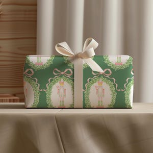 Op de afbeelding: Een rechthoekige geschenkdoos verpakt in groen papier met een herhalend patroon van notenkrakers in witte ovale kaders, omringd door groene kransen en roze linten, afgewerkt met een crèmekleurig satijnen lint gestrikt in een strik.