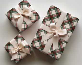 May include: Three rectangular gift boxes wrapped in cream, dark green, and red plaid paper. Each box is tied with a smooth, cream-colored satin ribbon arranged in a decorative bow centered on the top surface of the wrapping paper.