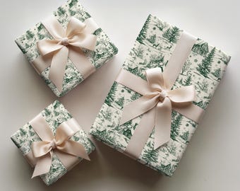 May include: Three rectangular gift boxes wrapped in white paper with a dark green winter forest scene featuring trees, cabins, and deer. Each box is tied with a smooth, pale pink satin ribbon arranged in a decorative bow centered on the top.
