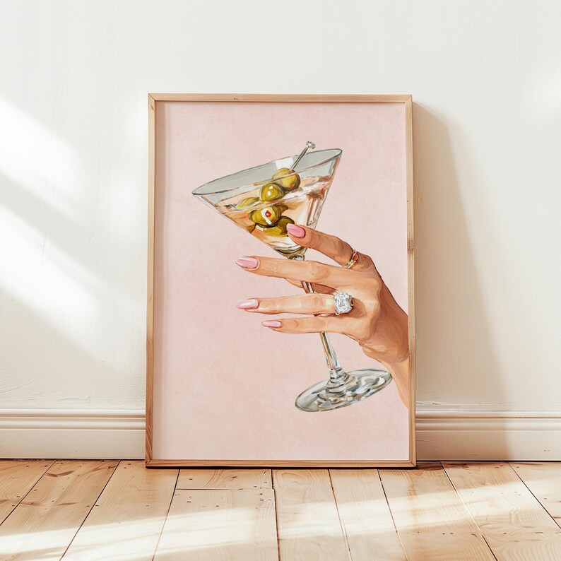 Op de afbeelding: Een ingelijste print met een hand die een martini-glas met olijven vasthoudt. De hand, met roze nagellak en ringen, staat tegen een roze achtergrond. Het kunstwerk straalt elegantie uit.