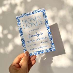 Può includere: Un biglietto d'invito rettangolare con un bordo a motivo di piastrelle ad acquerello blu e bianco. Il testo recita: HONEY HONEY, YOU'RE INVITED TO, Emily's, BRIDAL SHOWER, JULY 5-8TH, 1PM-4PM, Malibu, California. Il biglietto ha uno sfondo bianco.