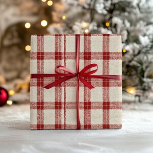 K&ouml;nnte beinhalten: Ein quadratischer Geschenkkarton, verpackt in cremefarbenem Papier mit einem roten Karomuster. Die Box ist mit einem roten Satinband und einer Schleife gebunden. Der Hintergrund zeigt einen verschwommenen Weihnachtsbaum und Lichter.