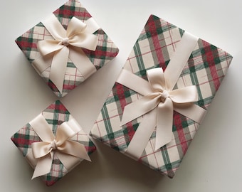 May include: Three rectangular gift boxes wrapped in cream-colored paper featuring a red and green plaid pattern. Each box is tied with a smooth, cream-colored satin ribbon arranged in a decorative bow centered on the top surface of the packages.