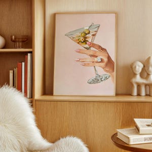 Op de afbeelding: Een ingelijste print met een hand die een martini-glas met olijven vasthoudt. De hand is versierd met ringen en heeft roze nagellak. De achtergrond is zachtroze en de print wordt gepresenteerd in een houten lijst.
