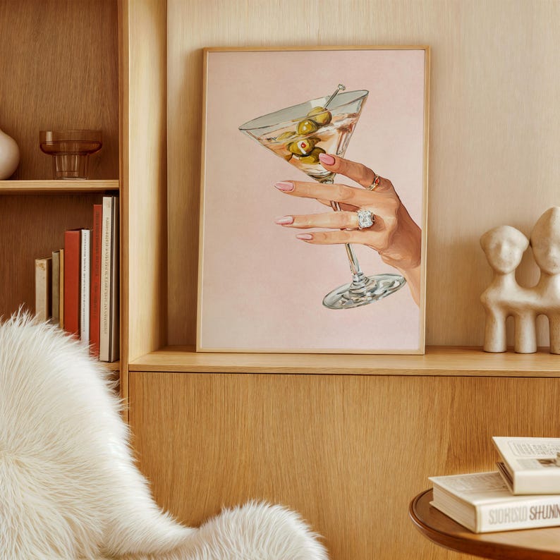 Op de afbeelding: Een ingelijste print met een hand die een martini-glas met olijven vasthoudt. De hand is versierd met ringen en heeft roze nagellak. De achtergrond is zachtroze en de print wordt gepresenteerd in een houten lijst.