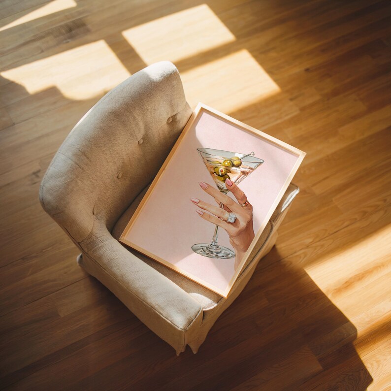 Op de afbeelding: Een ingelijste print met een martini-glas met olijven, vastgehouden door een hand met een ring en verzorgde nagels. Het kunstwerk heeft een roze achtergrond en een licht houten frame. De print rust op een beige fauteuil.