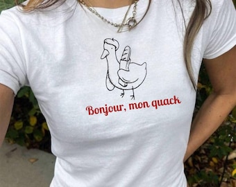 Puede incluir: Camiseta blanca con una ilustración en blanco y negro de un pato con un gorro y sosteniendo una baguette. El texto "Bonjour, mon quack" está impreso en rojo debajo de la ilustración.