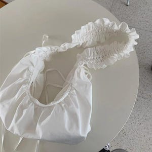 May include: A white, gathered fabric shoulder bag with a ruffled strap. The bag has a rounded shape and drawstring closure. The strap is adorned with a delicate ruffle detail, adding a touch of elegance to the design.
