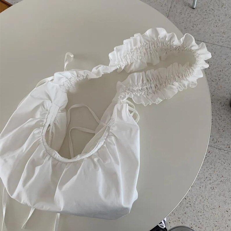 May include: A white, gathered fabric shoulder bag with a ruffled strap. The bag has a rounded shape and drawstring closure. The strap is adorned with a delicate ruffle detail, adding a touch of elegance to the design.