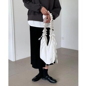 May include: A white tote bag with ruffled handles is held by a person. The bag has a simple, minimalist design. The person is wearing a black sweater, a white shirt, and a black skirt. Black shoes complete the outfit.