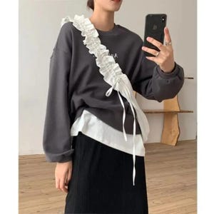 May include: A dark gray sweatshirt with a white ruffled accent cascading down the front. The sweatshirt is layered over a white top and paired with a black skirt. The image shows a casual, layered outfit.