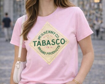 May include: A light pink t-shirt featuring the Tabasco brand logo in a diamond shape. The logo is in green and yellow. The shirt is paired with blue jeans and a white purse.