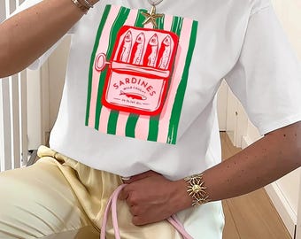 May include: White t-shirt featuring a sardine can graphic with the text "SARDINES WILD CAUGHT". The graphic is set against a pink and green striped background. The shirt is paired with light yellow pants.