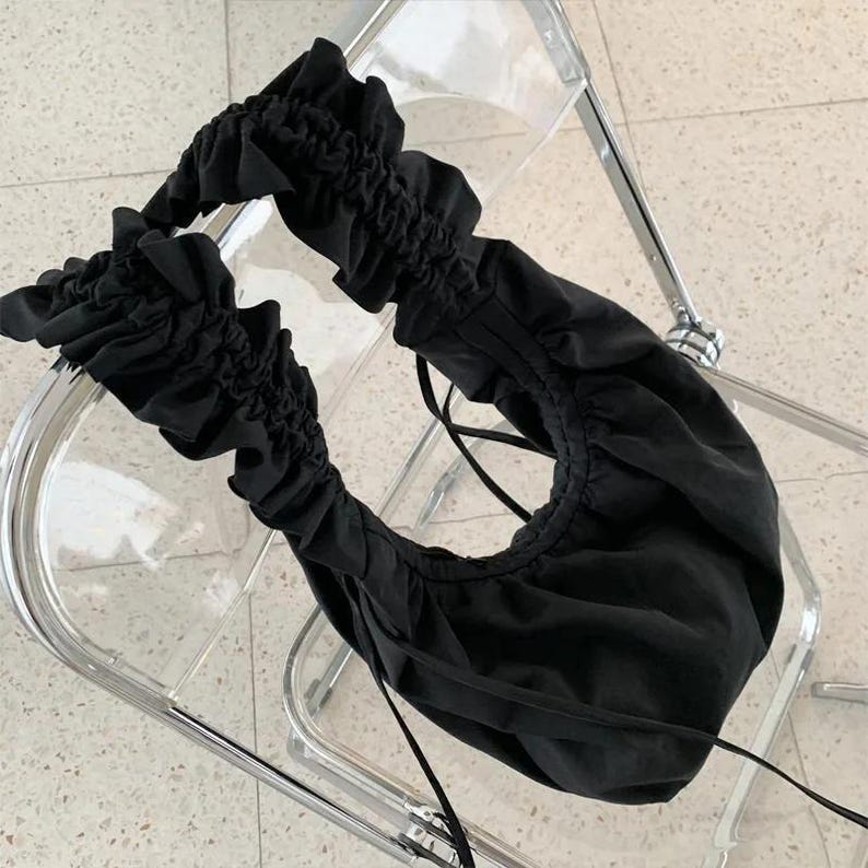 May include: Black off-the-shoulder top with ruffled sleeves and neckline. The top is made of a soft fabric and features a drawstring for an adjustable fit. The top is shown on a clear chair.