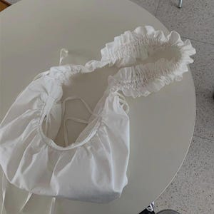 May include: A white, gathered fabric shoulder bag with a ruffled top edge. The bag has a rounded shape and features tie closures. The bag is made of a soft, lightweight material.