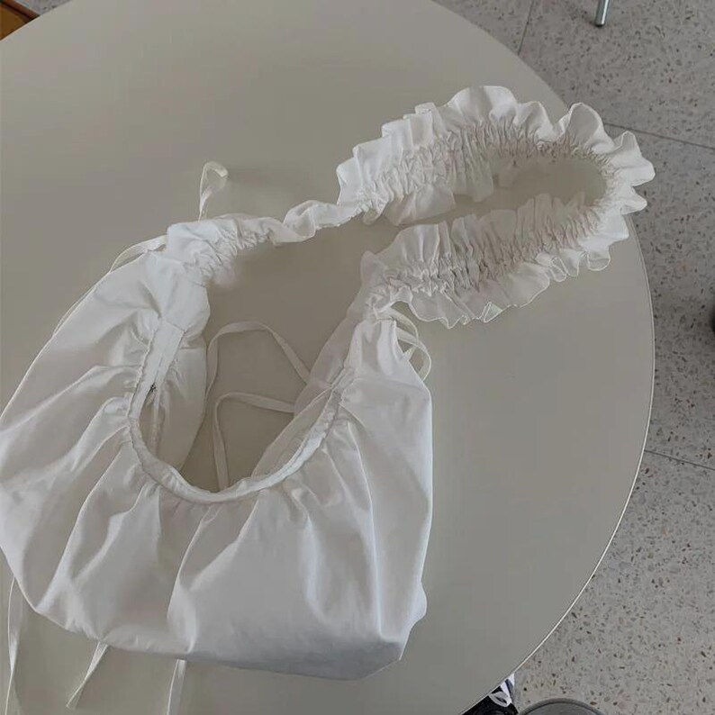 May include: A white, gathered fabric shoulder bag with a ruffled top edge. The bag has a rounded shape and features tie closures. The bag is made of a soft, lightweight material.