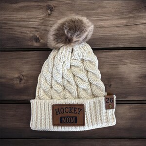 Puede incluir: Gorro de punto blanco con un pomp&oacute;n de piel sint&eacute;tica marr&oacute;n. El gorro tiene un parche de cuero marr&oacute;n que dice "HOCKEY MOM" y una etiqueta de cuero marr&oacute;n con el n&uacute;mero "26" en &eacute;l.