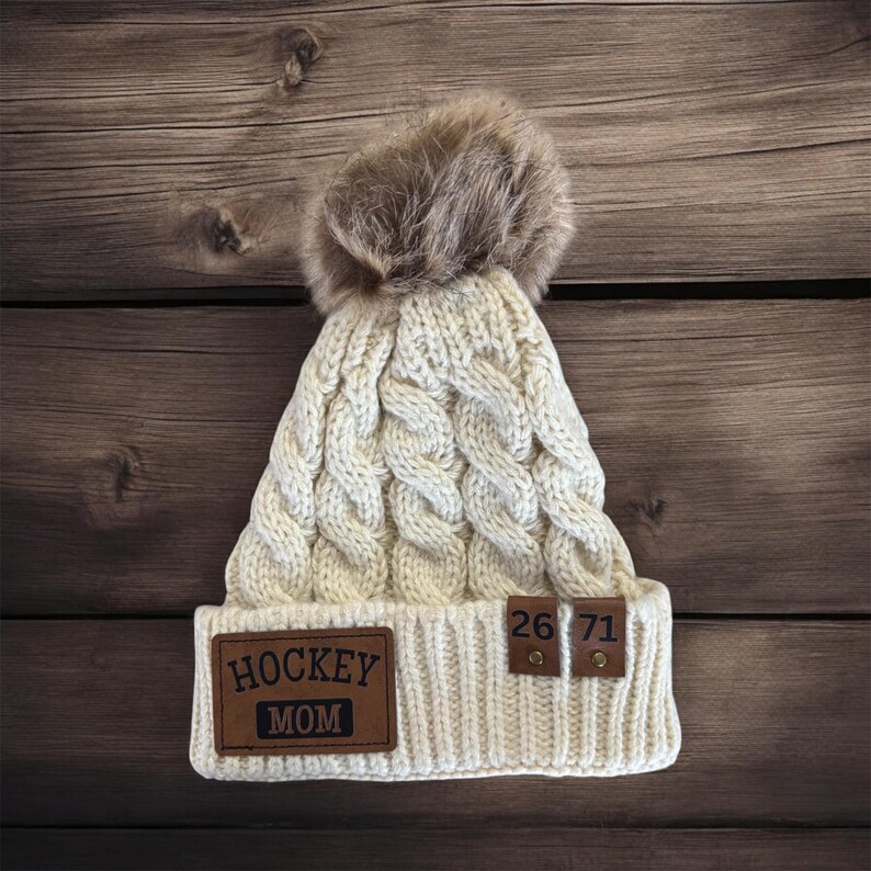 Puede incluir: Gorro blanco de punto con un pomp&oacute;n de piel sint&eacute;tica marr&oacute;n. El gorro tiene un parche de cuero marr&oacute;n que dice "HOCKEY MOM" y dos parches de cuero marr&oacute;n m&aacute;s peque&ntilde;os con los n&uacute;meros "26" y "71".