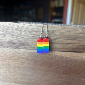 May include: A pair of earrings made from colorful toy blocks, arranged in a rainbow pattern. The earrings feature red, orange, yellow, green, blue, and purple blocks, with silver-colored ear wires. The earrings are displayed on a wooden surface.