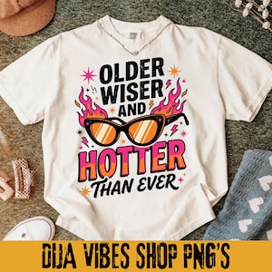 May include: White t-shirt with the text "OLDER WISER AND HOTTER THAN EVER" in a colorful design featuring sunglasses and flames. The shirt has a crew neck and short sleeves. The bottom of the image has the text "DIJA VIBES SHOP PNG'S".