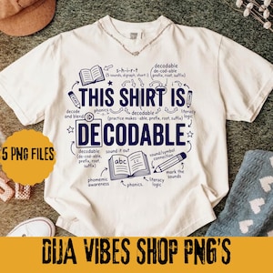 Op de afbeelding: Een crèmekleurig t-shirt met korte mouwen en donkerblauwe tekst en afbeeldingen. Het ontwerp bevat de woorden THIS SHIRT IS DECODABLE, omringd door illustraties van een open boek, een potlood en diverse educatieve termen zoals phonics.