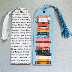 May include: Two decorative bookmarks. One is white with a tassel and a list of literary terms. The other is light blue with a stack of books, a typewriter, and flowers. The blue bookmark has a blue tassel.