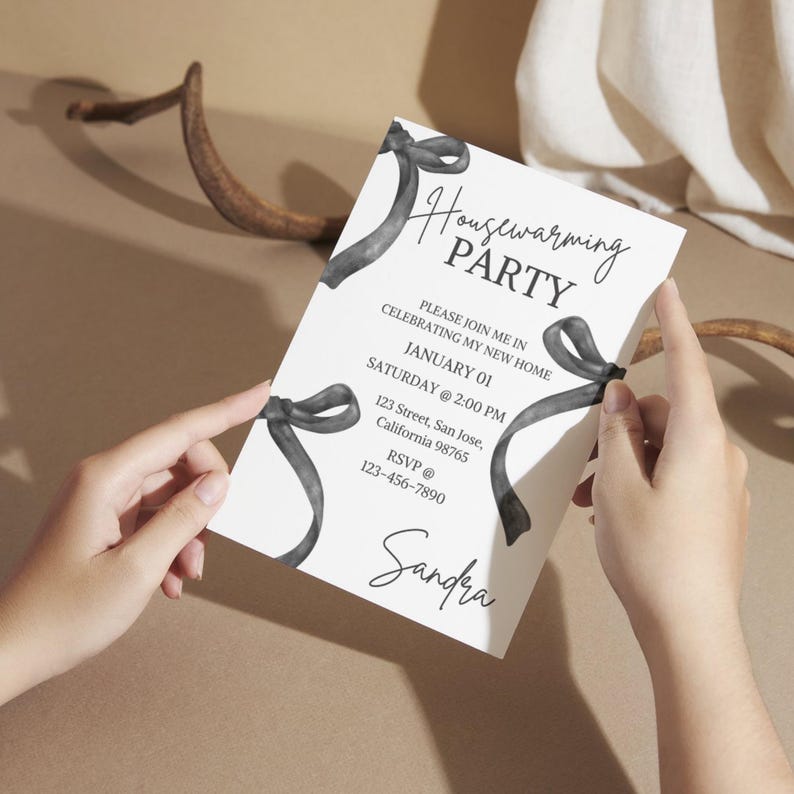 May include: A white invitation card with black watercolor ribbons and the text "Housewarming Party". The invitation includes the date, time, address, and RSVP information for a housewarming party. The name "Sandra" is written at the bottom of the card.