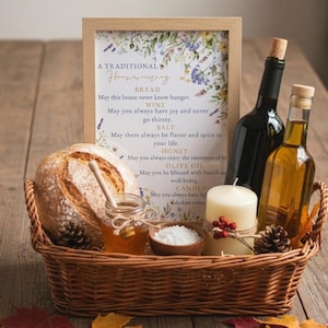 May include: A housewarming gift basket with a loaf of bread, honey, salt, olive oil, a candle, and two bottles of wine. A framed sign with a floral design and text is also included. The basket is made of woven wicker.