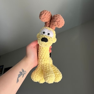 May include: A yellow crocheted dog toy with brown ears and black eyes. The toy is being held by a hand with a fish tattoo.